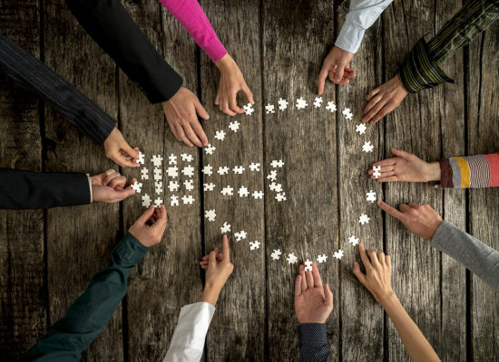 Teamwork and cooperation concept - group of twelve people, male and female, assembling a light bulb shape with blank puzzle pieces on a rustic textured desk, top view.