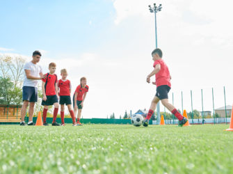Side view portrait of junior football team training outdoors  with focus on  red haired boy leading ball between orange cones, copy space